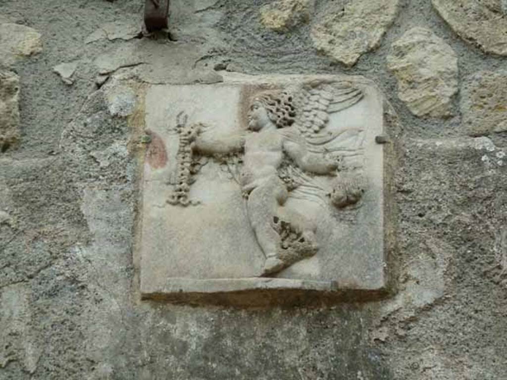 III.3 Herculaneum. May 2010. Detail of marble relief of cupid on wall above the pool.
The cupid is carrying in his hand, according to Jashemski, a purse filled with money.
See Jashemski, W. F., 1993. The Gardens of Pompeii, Volume II: Appendices. New York: Caratzas. (p.259)